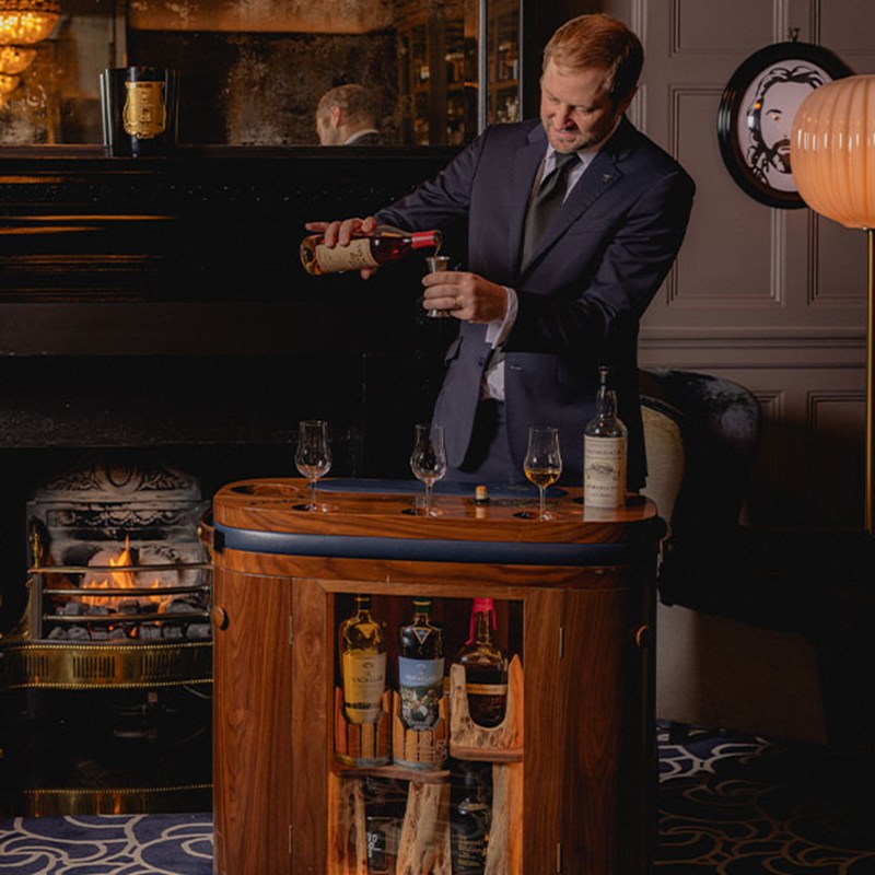 A bartender in a refined setting pouring a select spirit from a bottle, with a collection of whisky glasses arranged on a luxurious wooden cart