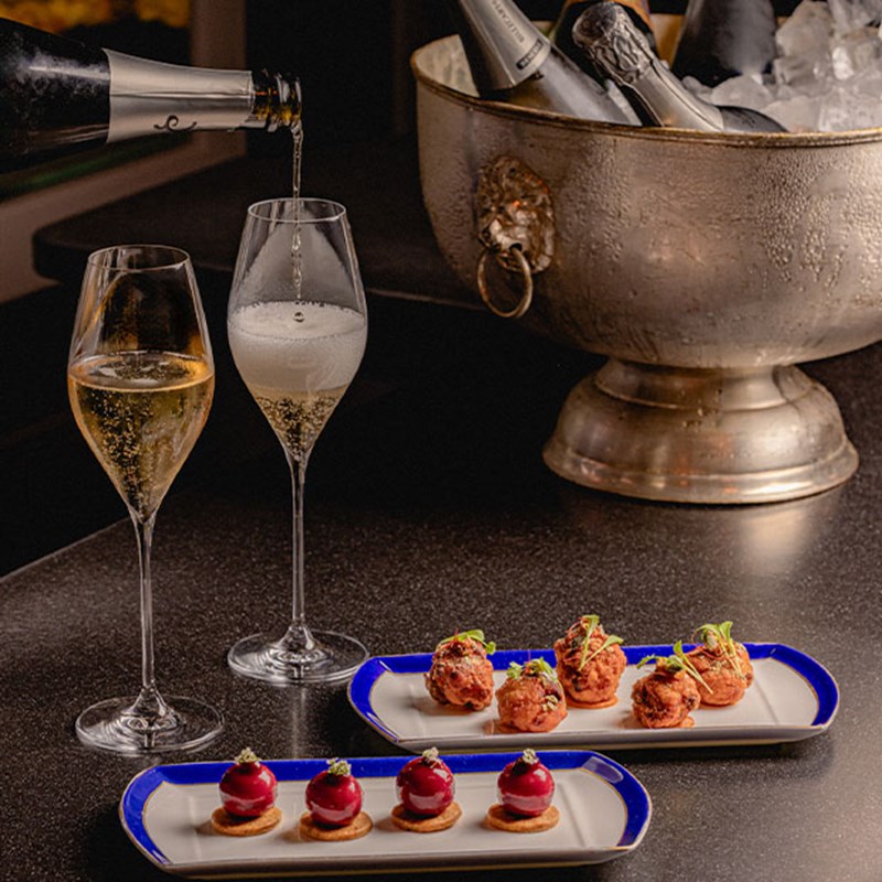 Two champagne flutes being filled alongside two plates of artfully presented canapés, set against a silver ice bucket filled with champagne bottles
