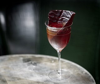 connaught bar burgundy cocktail in tall thin stemmed glass on a glass table with a dark green background