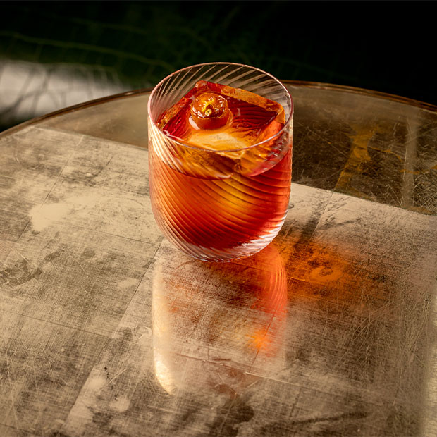 Amber cocktail in a ribbed glass with a large square ice cube on a reflective brass table.