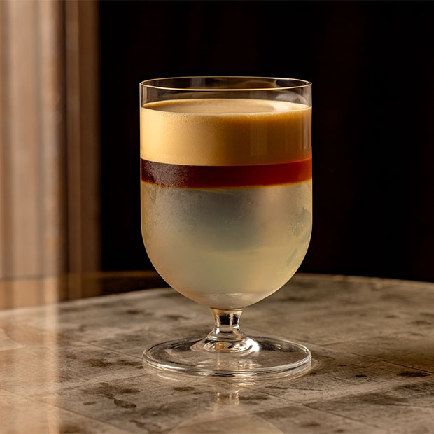 Layered cocktail in a stemmed glass with pale base, dark middle band and foamy top on a marble table.