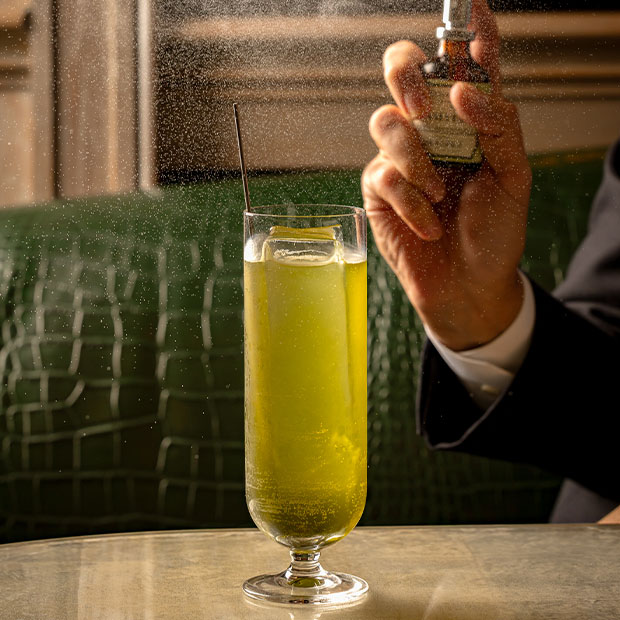 Tall green cocktail with ice as a hand sprays mist over the glass on a reflective table.