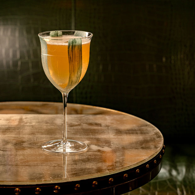 Golden cocktail in a stemmed glass with two green brushstroke garnishes on a brass table