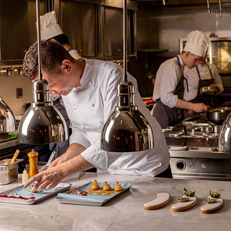 A chef in a white jacket adds finishing touches to plated appetisers under heat lamps in a professional kitchen, while other chefs work in the background.