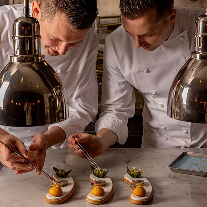 Two chefs in white uniforms use tweezers to meticulously plate garnished appetisers on curved ceramic dishes beneath stainless steel heat lamps.