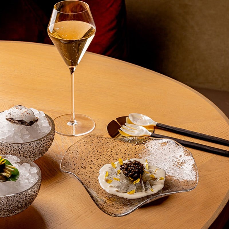 Close-up of an elegant caviar and cream-based seafood dish artfully arranged on a speckled glass plate, flanked by ice-filled bowls with garnished oysters.