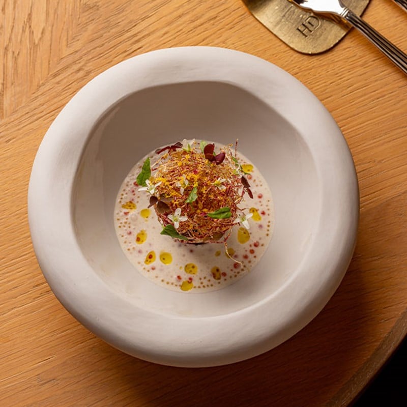 An intricately plated dish in a sculptural white bowl, featuring a central garnish of micro herbs and edible flowers over a creamy, dotted sauce.