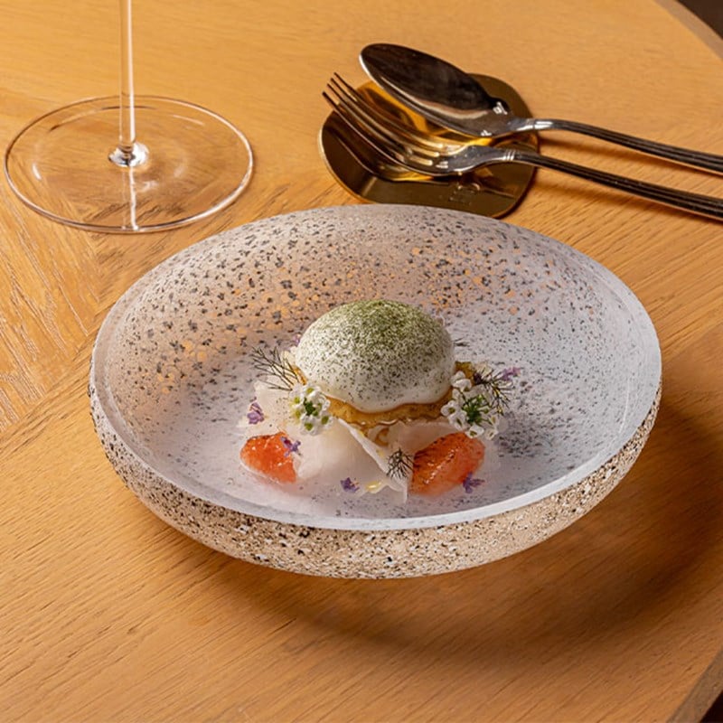 A textured glass bowl holding a delicately composed seafood and citrus dish, topped with a green mousse dome and surrounded by edible flowers.