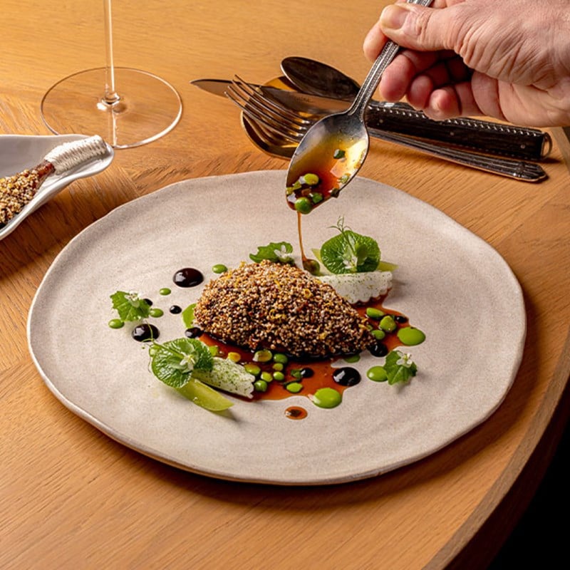 A hand pouring vibrant sauce over a dish encrusted in seeds, accompanied by green vegetable elements and colourful drizzles on a neutral-toned plate.