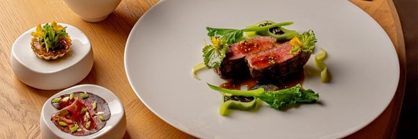 A fine dining presentation featuring sliced beef garnished with greens and edible flowers on a large white plate, with two small appetisers on elegant side dishes.