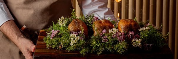 A chef presents three golden roast birds on a wooden board, garnished with fresh herbs and pink and white flowers, with a flame rising behind the centre bird.