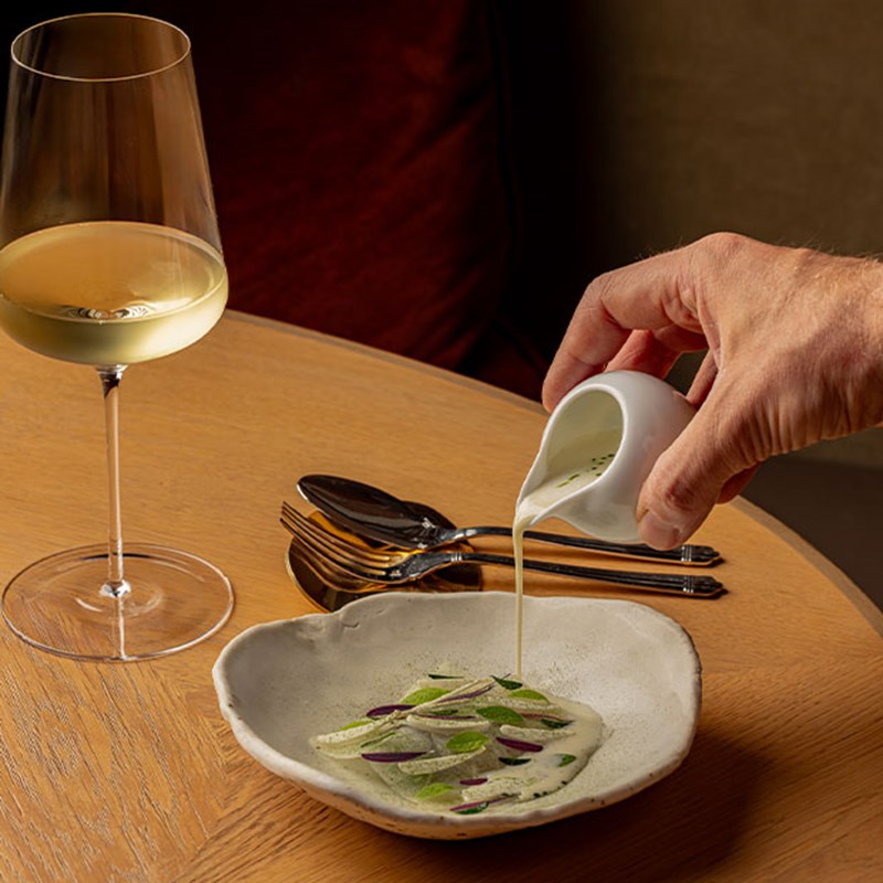 Hand pouring creamy soup into a bowl garnished with thinly sliced vegetables and herbs, with a glass of white wine beside it.