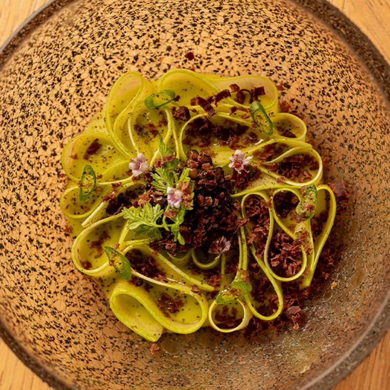 Artistic dish of zucchini ribbons arranged in a floral pattern, topped with herbs, edible flowers, and crispy garnish.