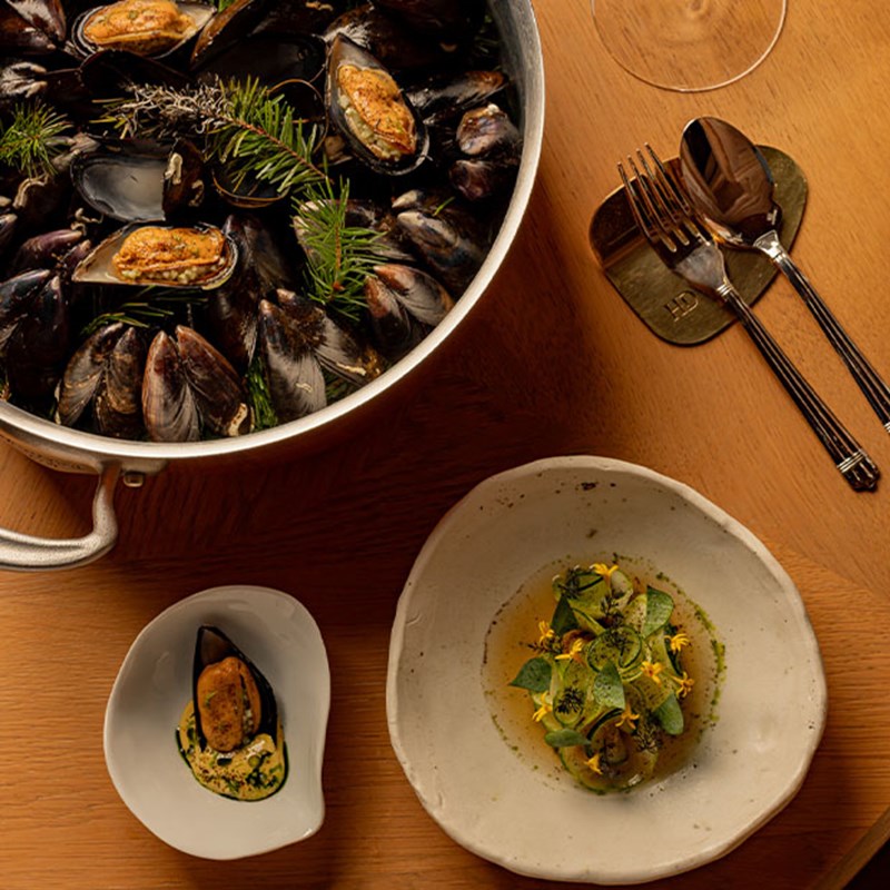 Large silver pot of mussels garnished with herbs, served alongside a small mussel appetiser and a plated dish with edible flowers and greens.