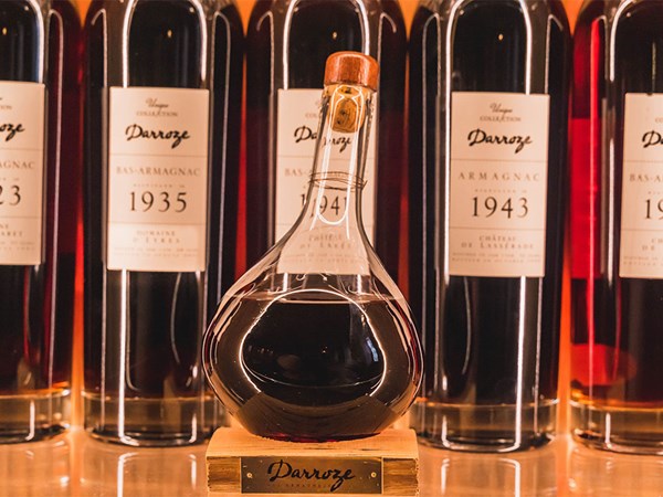 A glass decanter of dark Armagnac displayed in front of vintage Armagnac bottles from the 1930s and 1940s.