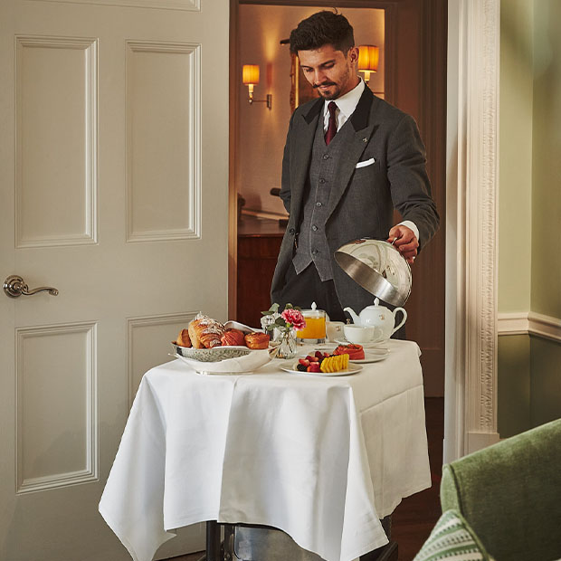 Smartly dressed hotel staff member in a tailored suit presenting a breakfast service in-room, with a white tablecloth trolley laid with pastries, tea, juice, and fresh fruit.