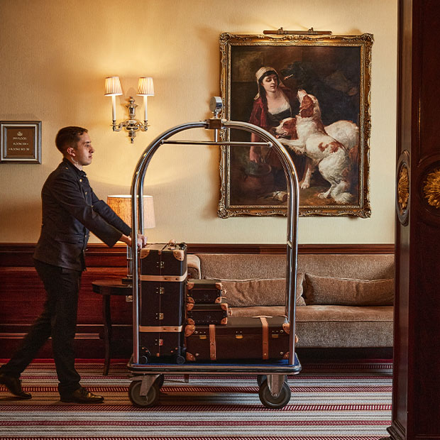 Hotel porter in uniform pushing a luggage trolley stacked with classic leather suitcases through an elegant corridor, beneath a framed painting of a woman and two dogs.