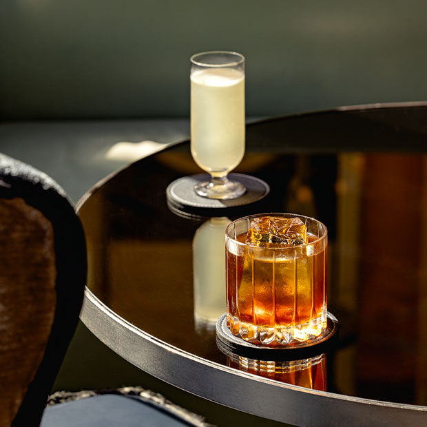 Two cocktails on a black lacquered table — one dark amber drink with ice in a tumbler and one pale yellow drink in a tall flute — bathed in natural light.