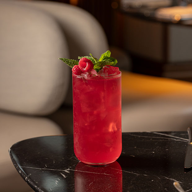 Vibrant red cocktail served in a tall glass with crushed ice, garnished with fresh raspberries and mint, placed on a black marble table.