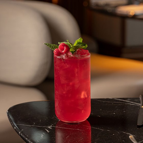 Vibrant red cocktail served in a tall glass with crushed ice, garnished with fresh raspberries and mint, placed on a black marble table.