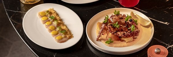 Dishes of grilled octopus with sauce and seared scallop crudo with yellow dressing, set on a black marble table with cocktails in the background.