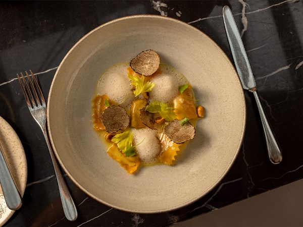 A ceramic bowl of filled pasta topped with foam, black truffle slices, nuts and small green herb leaves, placed on a dark marble table with cutlery beside it.