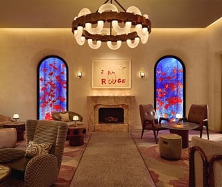 A warm, art-filled lounge featuring a central fireplace beneath a statement chandelier, flanked by stained glass windows with vibrant red floral motifs and contemporary seating arranged throughout.
