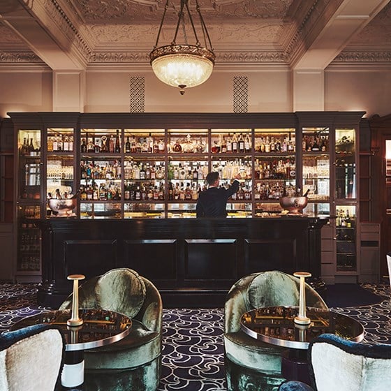 View of the luxurious interior of the Coburg Bar, in The Connaught, and a barman selecting a drink from the shelf.