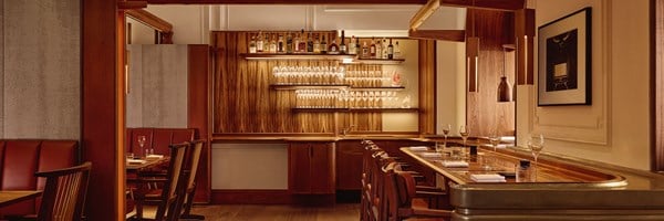 The Connaught Grill’s elegant bar area, showcasing high stools, a polished wood counter, and shelves of gleaming glassware and premium spirits set against a striped wood backdrop.