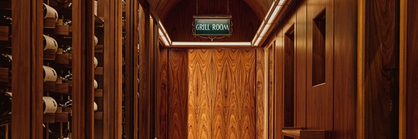 A warmly lit corridor leading to The Connaught Grill, lined with wine storage on either side and finished in rich wood paneling, with a vintage-style ‘Grill Room’ sign above the arched entrance.