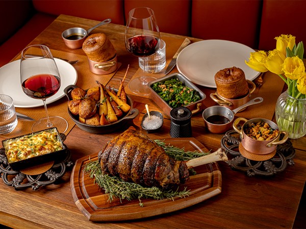 Wooden table set for Sunday lunch with a roasted leg of lamb, roasted vegetables, gratin, glasses of red wine, assorted side dishes, and a vase of yellow tulips.
