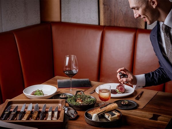 A suited server sprays a fine mist over a plated meat dish at a restaurant table set with wine, bread, knives, and gourmet plates.