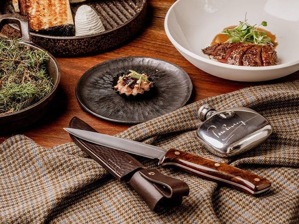 A close-up of a restaurant table showing a plated meat dish, a tartlet, artisan bread, a silver hip flask, and a knife on a checked wool cloth.