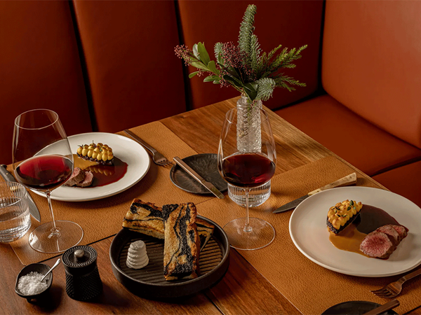 Elegant restaurant table with two plated meat dishes in rich sauce, red wine glasses, artisan bread, and floral centerpiece.