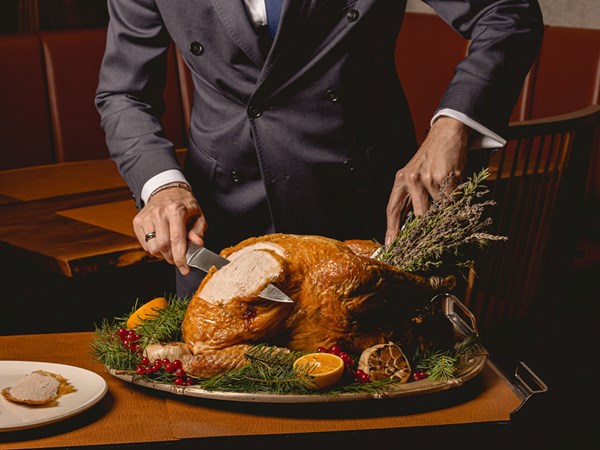 A person in a tailored suit carves a golden roast turkey on a silver platter garnished with herbs, cranberries, orange slices and roasted garlic, with one slice placed on a nearby plate.