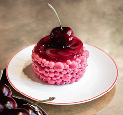 A glossy cherry-topped pâtisserie with a textured pink glaze sits on a white plate with red trim, with fresh cherries in a silver dish beside it on a reflective surface.