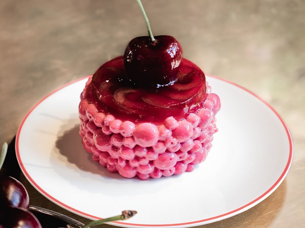 Close-up of glossy cherry-themed pastries with pink glazed coating and a fresh cherry on top, arranged on a marble surface.