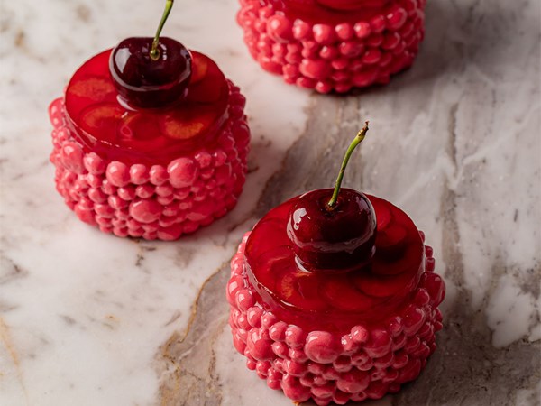 Close-up of glossy cherry-themed pastries with pink glazed coating and a fresh cherry on top, arranged on a marble surface.