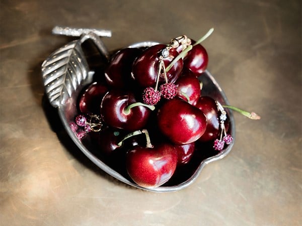 Fresh cherries arranged in a silver dish, accented with sparkling jewellery designed to mimic cherry stems, on a bronze-toned surface.