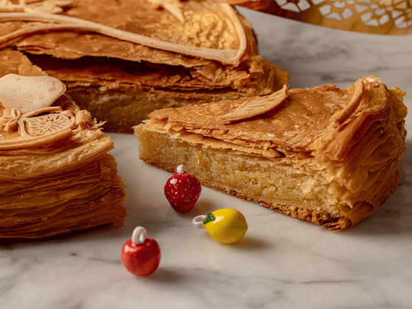 Slices of golden galette des rois with flaky pastry layers, shown with colourful ceramic fèves on a marble surface.