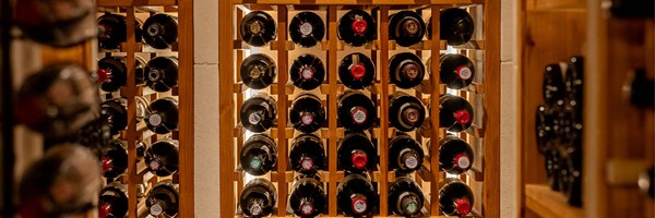 A warmly lit wooden wine rack filled with horizontally stored bottles, viewed from the base of the bottles with visible labels and coloured caps.