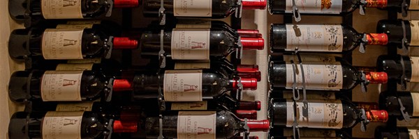 A warmly lit wooden wine rack filled with horizontally stored bottles, viewed from the base of the bottles with visible labels and coloured caps.