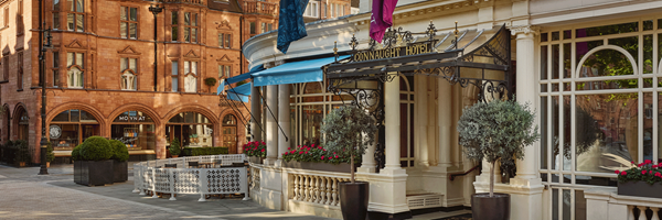 Exterior view of The Connaught Hotel in Mayfair, with ornate signage, potted trees, and the red brick Moynat storefront opposite.
