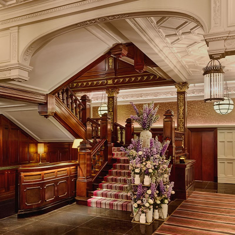 Grand wooden staircase inside The Connaught, adorned with purple floral arrangement, under ornate ceiling mouldings and ambient lighting