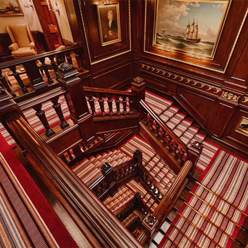 Multi-level staircase with striped carpet and oil paintings in a dark wood interior, viewed from above at The Connaught.
