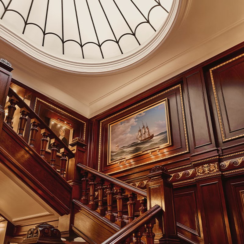 View looking up at framed ship painting and circular skylight, surrounded by dark wood panelling and gold accents.
