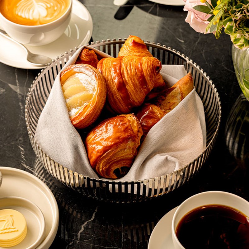 ChatGPT said: A basket of assorted golden pastries, including croissants and fruit danishes, lined with a cloth napkin on a marble table set with coffee, butter, and a cappuccino.