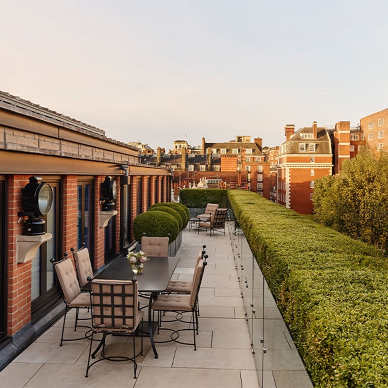 Elegant rooftop terrace with cushioned seating and dining table, overlooking historic Mayfair rooftops and manicured hedges at golden hour.