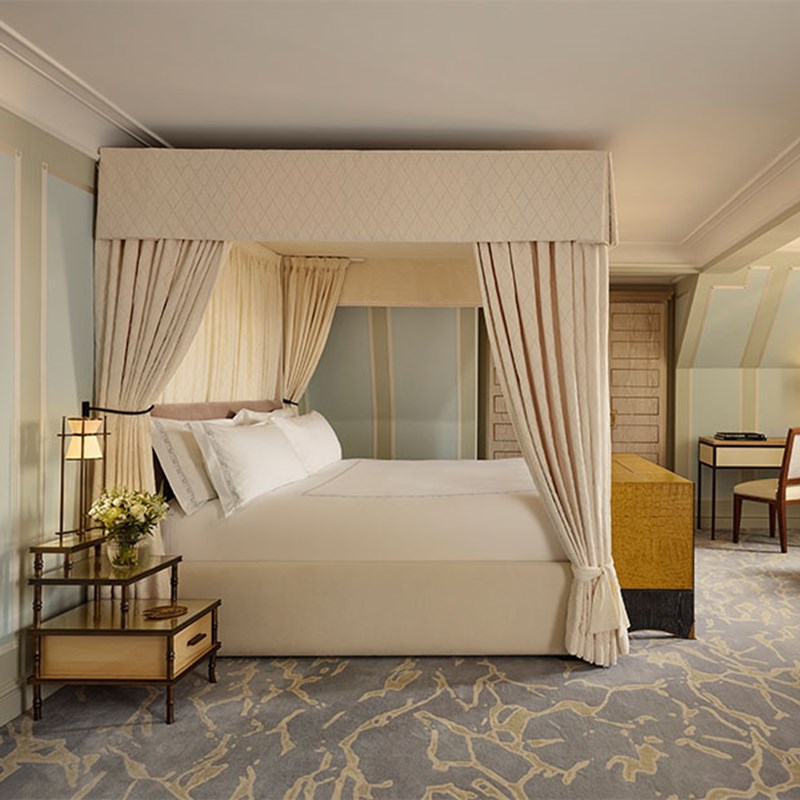 Elegant guest room with a cream four-poster bed, patterned carpet, bedside flowers, and light wood furnishings in soft lighting.