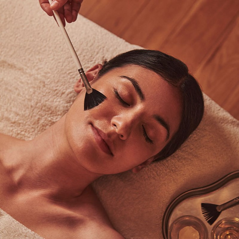 Woman receiving a facial with a fan brush, lying on a towel-covered table with a tray of tools nearby.